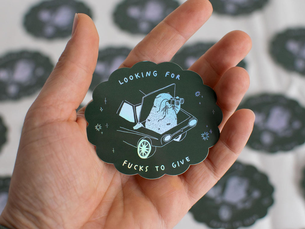 Looking for fucks to give 2.0 | Holographic Cat Sticker of a cat sitting in the trunk of a car looking for f***s to give through binoculars